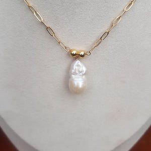 Puede incluir: Un collar de cadena dorada con un colgante de perla blanca grande e irregular. La cadena tiene un dise&ntilde;o de eslabones alargados. Dos peque&ntilde;as cuentas doradas est&aacute;n situadas encima de la perla. El collar se muestra sobre un fondo neutro.