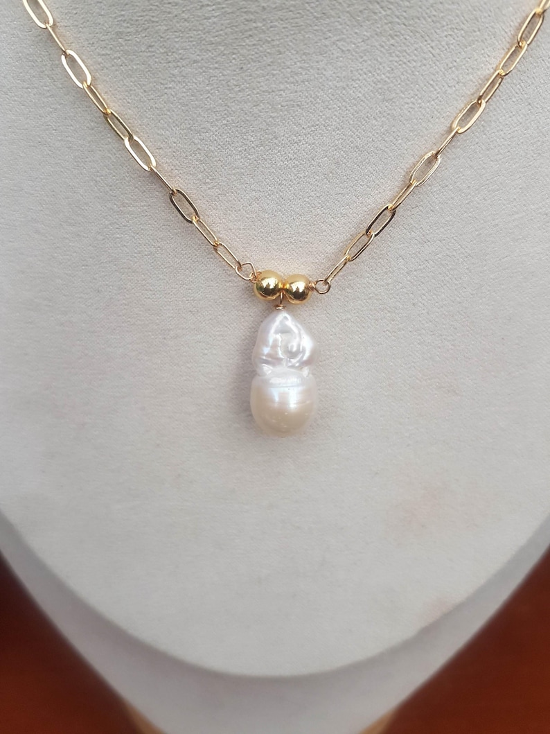Puede incluir: Un collar de cadena dorada con un colgante de perla blanca grande e irregular. La cadena tiene un dise&ntilde;o de eslabones alargados. Dos peque&ntilde;as cuentas doradas est&aacute;n situadas encima de la perla. El collar se muestra sobre un fondo neutro.