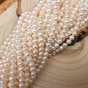 Puede incluir: Una hebra de perlas de agua dulce blancas, dispuestas en una pila sobre una superficie de madera. Las perlas son redondas y lisas, con un ligero brillo iridiscente.