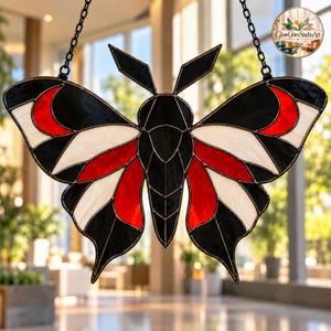 Puede incluir: Un atrapasoles de vidrieras con forma de polilla, con un diseño simétrico de segmentos de vidrio negro, blanco y rojo unidos por líneas de soldadura de metal oscuro. La polilla tiene dos antenas y dos cadenas metálicas negras para colgar.