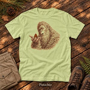 May include: A light green t-shirt featuring a detailed brown illustration of a hairy creature resembling a Bigfoot holding a small pinecone in its hand. The graphic is centered on the chest of the shirt, showcasing fine line work and shading.