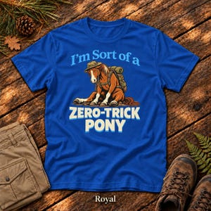 May include: A royal blue short-sleeve t-shirt featuring a graphic of a brown pony wearing a hat and a backpack. The text above the pony reads "I'm Sort of a" and the text below reads "ZERO-TRICK PONY" in bold white and light blue lettering.