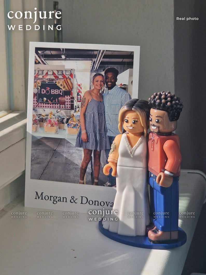 Op de afbeelding: Een aangepaste bruidstaarttopper met twee plastic figuren die een koppel voorstellen, naast een afgedrukte foto van het tweetal. De figuren zijn gestileerd als bouwsteenspeelgoed, met een figuur in een witte jurk en een figuur in een oranje shirt en blauwe broek. De foto bevat de tekst Morgan & Donovan.