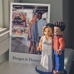 May include: A custom wedding cake topper featuring two plastic figures resembling a couple, standing next to a printed photograph of the pair. The figures are styled like building block toys, with the female figure in a white gown and the male figure in an orange shirt and blue pants. The photo includes the text Morgan & Donovan.
