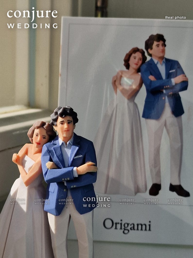 Op de afbeelding: Een 3D-geprint beeldje van een koppel, waarbij de persoon links een witte strapless jurk draagt en de persoon rechts een blauwe blazer, een lichte broek en een wit overhemd, weergegeven voor een bijpassende bedrukte kaart.