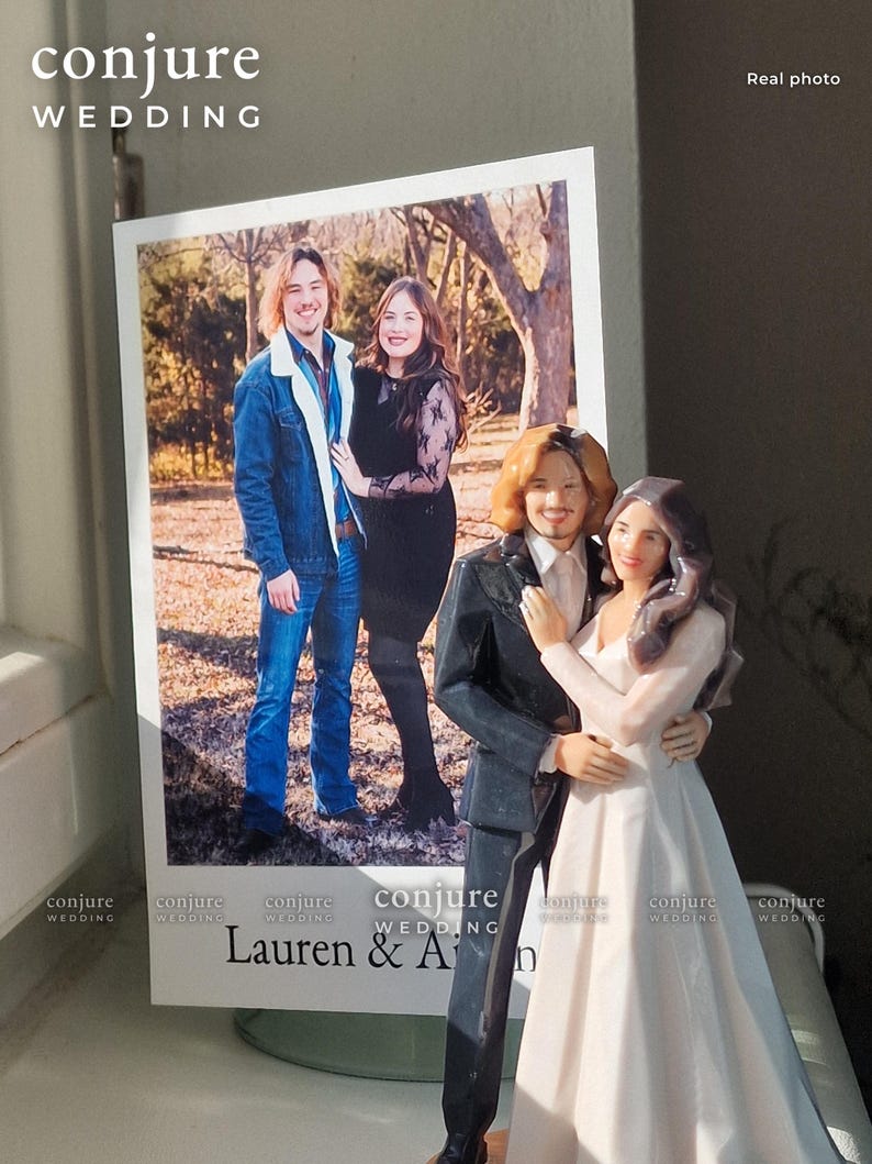 May include: A custom wedding cake topper figurine depicting a couple in formal attire, standing next to a printed photograph of the same couple in casual clothing. The text "conjure WEDDING" and "Lauren & An" are visible on the display.