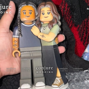 May include: A custom figurine set featuring two block-style figures standing together. One figure has braided hair, a dark blue shirt, and gray pants. The other has long brown hair, a light green shirt, and a long black skirt. Text reads conjure WEDDING.