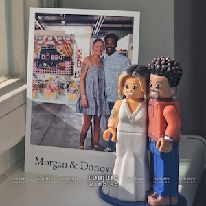 May include: A custom wedding cake topper featuring two plastic figures resembling a couple, standing next to a printed photograph of the pair. The figures are styled like building block toys, with the female figure in a white gown and the male figure in an orange shirt and blue pants. The photo includes the text Morgan & Donovan.