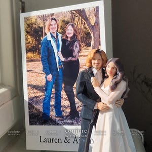 Op de afbeelding: Een op maat gemaakte bruidstaartfiguur die een koppel in formele kleding voorstelt, naast een afgedrukte foto van hetzelfde koppel in vrijetijdskleding. De tekst "conjure WEDDING" en "Lauren & An" is zichtbaar op de display.