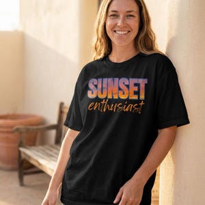 May include: A black short-sleeved t-shirt featuring the text SUNSET in bold, block letters filled with a sunset gradient of orange, purple, and yellow, followed by the word enthusiast in a cursive orange script font centred on the chest.