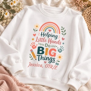 May include: A white crewneck sweatshirt featuring a colorful graphic with a rainbow, flowers, handprints, and a pencil. The text reads: Helping Little Hands Do BIG Things, Jessica, OTR/L. The design includes small hearts and stars.