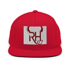 May include: A bright red baseball cap featuring a rectangular patch on the front. The patch is light grey with a quilted texture and displays the letters RH in a dark red font, with the R and H stylised to include devil horns and a tail.
