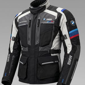 Puede incluir: Una chaqueta de moto negra, gris y blanca con el logotipo de BMW en el pecho y la manga. La chaqueta incluye múltiples bolsillos con cremallera, un cuello alto, correas laterales ajustables y rayas decorativas azules y rojas en el brazo.