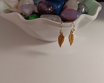 May include: A pair of gold-colored leaf earrings. The earrings are hanging in front of a white bowl filled with colorful stones. The earrings have a hook closure.
