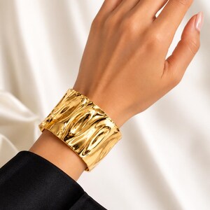 May include: A wide, polished gold-tone cuff bracelet featuring a distinct, irregular, and undulating surface texture. The metal has a high-shine, reflective finish that catches the light, creating deep shadows and bright highlights across the curved form.