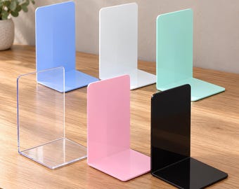 May include: A set of six L-shaped bookends made of smooth acrylic. The collection includes one clear, one white, one black, one light blue, one pastel pink, and one mint green bookend, each featuring a flat vertical panel and a base.
