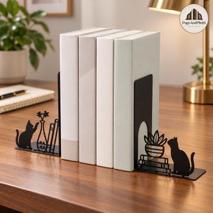 May include: A pair of black metal bookends featuring cat silhouettes. The left bookend displays a cat reaching towards a vase with flowers and books. The right bookend shows a cat sitting next to a potted plant on a stack of books.