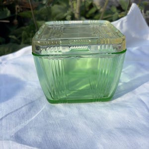 Op de afbeelding: Een vierkante, lichtgroene glazen container met bijpassend deksel. Het stuk heeft een geometrisch, geperst patroon met verticale lijnen langs de zijkanten en een rasterontwerp op de bovenkant van het deksel. Het glas heeft een doorschijnende tint.