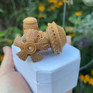 May include: A light tan, carved plastic sewing thimble featuring a decorative, star-shaped top with radiating ridges and a perforated, tiered base. The thimble is positioned on top of a white, hexagonal-shaped box, showcasing intricate detailing.