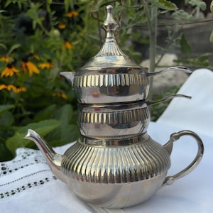 May include: A polished silver-tone metal double teapot stack featuring a large base pot with a curved spout and handle, topped by a smaller secondary pot with a matching lid. Both sections display vertical fluted detailing along their bodies.