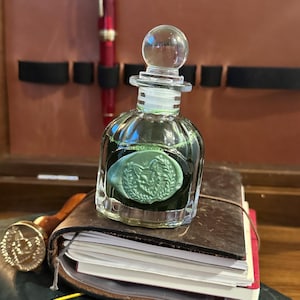 May include: A clear glass ink bottle with a rounded stopper, filled with dark green ink. The bottle features a pale green wax seal on the front, embossed with a detailed fox head design surrounded by a laurel wreath, resting on a small leather notebook.