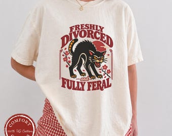 May include: An off-white short-sleeve t-shirt featuring a graphic of a black cat with arched back and bared teeth. The text reads FRESHLY DIVORCED and FULLY FERAL in red lettering, surrounded by red cherry blossom branches and a red sun circle.