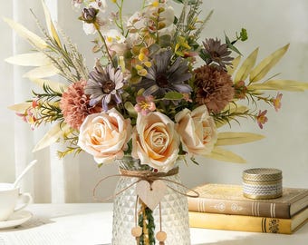 May include: A floral arrangement featuring peach roses, mauve chrysanthemums, purple daisies, and white orchids in a clear, diamond-patterned glass vase. The vase is decorated with a twine bow and a small wooden heart ornament.