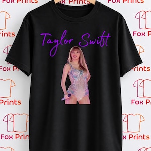 May include: A black short-sleeved t-shirt featuring a graphic of Taylor Swift wearing a sparkling, light-coloured bodysuit. The name Taylor Swift is printed in a purple script font above the image. The shirt is displayed on a wooden hanger.