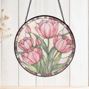 May include: A circular stained glass suncatcher featuring a bouquet of pink tulips with green stems and leaves. The piece is framed in a black metal border and hangs from a black metal chain. The glass has a textured, multi-coloured finish.