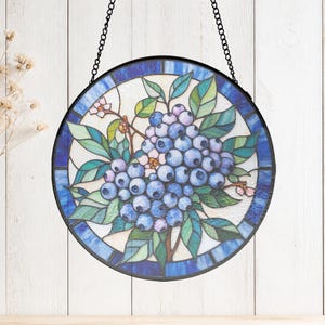 May include: A circular stained glass suncatcher featuring a cluster of blue berries and green leaves. The design is framed by a dark metal border and includes a black hanging chain. The glass pieces display various shades of blue, teal, and white.