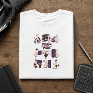 May include: A white t-shirt featuring a graphic print of various cartoon ducks engaged in computer programming tasks. The central text reads "Compile Then Panic" in a stylized font. The illustrations depict ducks with laptops, servers, and headsets.