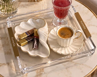 May include: A clear acrylic serving tray with gold-toned metal handles engraved with the name Kumelia. The tray holds a white shell-shaped dish containing three wrapped chocolates and a white fluted espresso cup and saucer with gold trim.