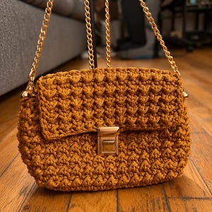 May include: A mustard yellow crochet handbag featuring a textured stitch pattern, a rectangular flap, and a gold-tone metal push-lock closure. The bag includes a matching gold-tone metal chain shoulder strap attached to the sides with gold hardware.