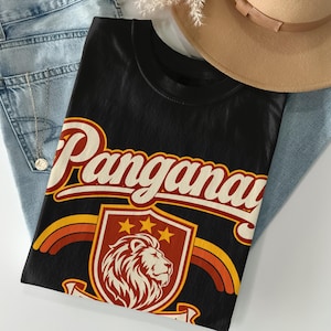 May include: A black t-shirt featuring the word Panganay in large white script with a red outline. Below the text is a shield emblem containing a lion head illustration, three stars, and curved orange and yellow stripes on either side.