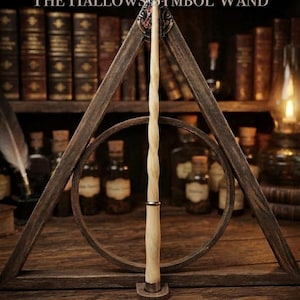 May include: A light-colored wooden wand with a spiral-carved shaft and a smooth handle, mounted vertically within a dark wooden triangular frame featuring a circular center. The text THE HALLOWS SYMBOL WAND is printed at the top of the image.