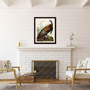 May include: A framed print of a detailed illustration of a wild turkey, with brown, gray, and white feathers, standing in a grassy field. The print is in a black frame and is hanging on a white brick wall above a fireplace.