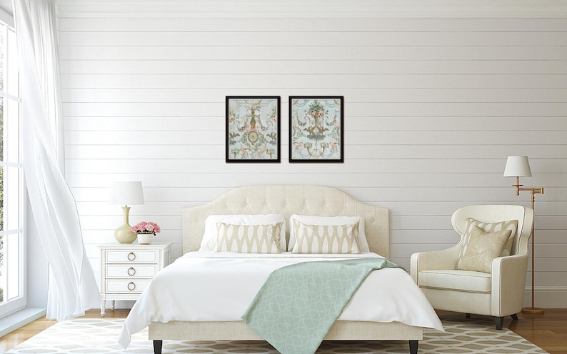 May include: A white upholstered bed with a light blue throw blanket and two framed prints of floral designs above the headboard. The bed is in a bedroom with white walls and a white wood floor. There is a white nightstand with a lamp and flowers on it to the left of the bed. A white armchair with a beige pillow is to the right of the bed.
