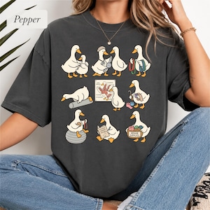 May include: A charcoal gray t-shirt featuring a graphic print of white ducks engaged in various physical therapy activities. The design includes ducks using exercise bands, a balance ball, and a basket labeled Rehab Supplies with text.