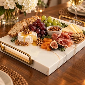 May include: A rectangular white marble serving tray with gold-toned metal handles on each side. The tray is arranged with an assortment of food items including sliced bread, red grapes, green grapes, almonds, cheese, walnuts, dried apricots, figs, and crackers.