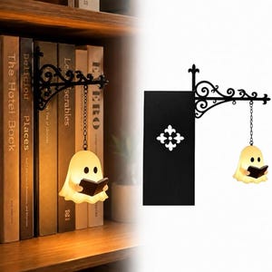 May include: A black metal wall-mounted bracket with ornate scrollwork, featuring a hanging light shaped like a small white ghost holding a black book. The bracket includes a vertical rectangular black panel with a white fleur-de-lis emblem.