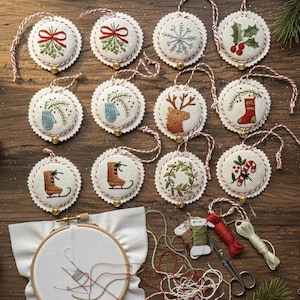 May include: Twelve round fabric ornaments featuring embroidered holiday designs including mistletoe, snowflakes, holly, mittens, reindeer, stockings, ice skates, and wreaths, each with a small gold bell and red and white hanging string.