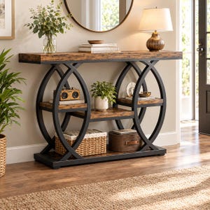 Può includere: Un tavolo console rettangolare con piano in finitura venatura del legno e due ripiani inferiori, sostenuto da telai in metallo nero con grandi accenti circolari laterali. Il tavolo ospita una lampada, libri, una piccola pianta e cesti.