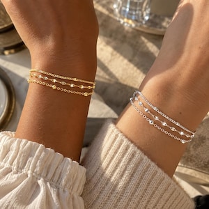 Könnte beinhalten: Ein Set aus zwei dreireihigen Armbändern, eines in Goldoptik und eines in Silberoptik. Jedes Armband besteht aus drei feinen Ketten: einer einfachen Gliederkette, einer Kette mit kleinen Metallperlen und einer Kette mit flachen, reflektierenden, rautenförmigen Gliedern.