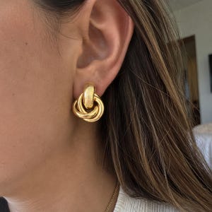 Puede incluir: Un pendiente de tono dorado pulido con un diseño de nudo liso y redondeado. El pendiente consta de un aro superior sólido conectado a una serie de bandas metálicas circulares entrelazadas que forman una forma de nudo continuo.