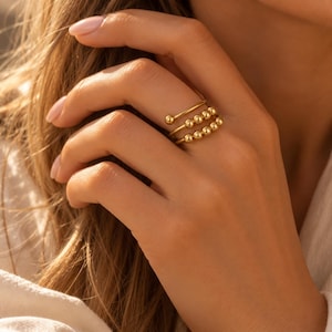 Puede incluir: Un anillo de metal en tono dorado con un diseño en espiral de varias capas. El anillo consta de tres bandas finas, con las dos bandas inferiores adornadas con una serie de pequeñas cuentas doradas, lisas y redondas, espaciadas uniformemente a lo largo del alambre.