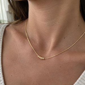 Puede incluir: Un collar de cadena en tono dorado con cinco pequeñas cuentas metálicas redondas centradas a lo largo de la delicada cadena de eslabones. El collar descansa sobre el cuello, mostrando un diseño sencillo con un acabado pulido en las cuentas y los eslabones.