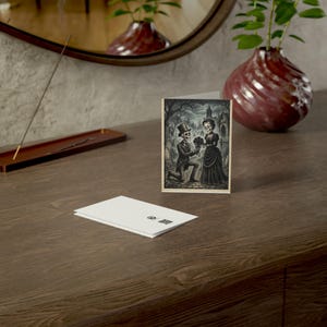 May include: A folded greeting card featuring a black and white illustration of a skeleton in a top hat kneeling to offer a bouquet of flowers to a skeleton in a dress, set in a graveyard with a gothic building in the background.