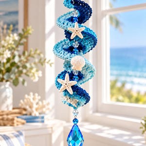 Könnte beinhalten: Ein hängendes Spiralornament aus gehäkeltem Garn in Marineblau, Königsblau und Hellblau. Die Spirale ist mit zwei weißen Seesternen, einer kleinen Muschel, klaren Glasperlen und einem großen, facettierten tropfenförmigen blauen Kristallanhänger verziert.