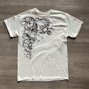 May include: A cream-coloured short-sleeve t-shirt featuring a large, detailed black and brown ink-style graphic print on the left side. The design depicts a skull surrounded by swirling, organic, smoke-like patterns that extend from the shoulder down.
