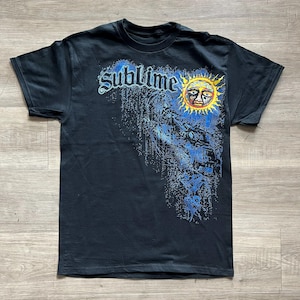 May include: A black short-sleeve t-shirt featuring the word Sublime in a stylized blue font across the chest. To the right of the text is a graphic of a yellow and orange sun with a face, surrounded by a blue textured pattern that fades downward.